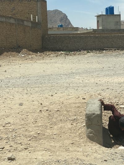The image depicts a dusty, vacant lot in Arghandab, Afghanistan. A person wearing dark clothing is crouched beside a rectangular concrete block. The surrounding terrain is gravelly and barren. Walls made of brick, possibly unfinished, enclose the lot. In the background, there are more low-lying structures and a mountain range under a hazy sky. Visible on the rooftops are blue water tanks.