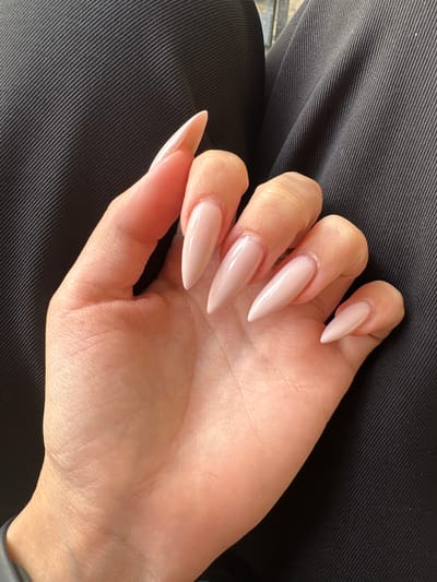 A close-up, overhead shot shows a person's left hand with stiletto-shaped, pale pink acrylic nails resting on a textured black fabric. The nails are uniformly polished and have a high-gloss finish. The fingers are slightly curled, with the palm facing upwards. The fabric appears to be a dark material with a subtle ribbed texture, possibly clothing or upholstery. The lighting suggests daylight, with soft shadows indicating an indoor or shaded outdoor setting. There are no visible signs of text in the image. The focus is entirely on the hand and nails, creating an intimate and detailed view of the subject's manicure. The overall impression is one of neatness and attention to personal grooming.