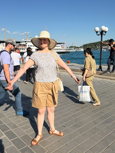 This image captures a sunny daytime scene on a paved walkway in Beyoğlu, Turkey, likely near the waterfront. A woman wearing a wide-brimmed hat, a floral top, and tan shorts stands prominently in the foreground, with her arms outstretched. She is holding hands with a man on her left, who is wearing a grey baseball cap, sunglasses, and blue jeans. Behind them, other individuals are walking, including a woman in tan clothing carrying a shopping bag and a man on the right holding a phone to his ear. Ferries and boats are visible on the blue water in the background, with trees and buildings on the distant shore. A lamppost with three globes stands to the right of the frame.