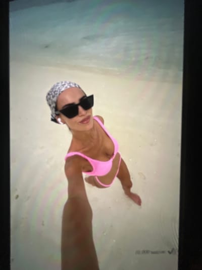 A woman is taking a selfie on a beach in Ravno pole, Bulgaria. She is wearing a pink bikini, black sunglasses, and a leopard-print headscarf. The water is shallow and clear, and the sand is white. The woman is smiling and looking at the camera. The overall scene is bright and sunny.