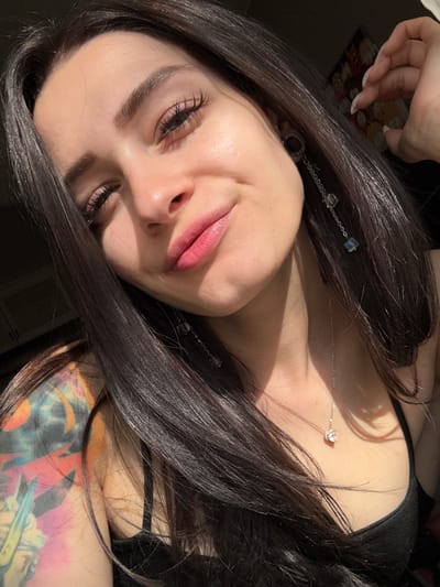 The image depicts a woman with long, dark brown hair, a light complexion, and visible tattoos on her left arm and shoulder. She is wearing a black top and has a silver necklace with a heart-shaped pendant. Her hair is adorned with silver chains and cube-shaped beads. She is positioned at an angle, looking upwards towards the camera, with a slight smile. The background is dark and underexposed. There is no indication of the location being Reutov, Russia, or any other specific location.