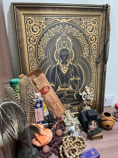 The image shows a shelf with various items related to spirituality and meditation. The central subject is a framed artwork featuring a mandala design with a depiction of what appears to be Buddha. Surrounding the artwork are several objects, including prayer beads, small figurines of deities or spiritual figures, incense sticks, feathers, and a small gourd-like object. The lighting suggests it's daytime indoors, with artificial light casting shadows. The overall mood is one of quiet contemplation and spiritual practice.
Visible text includes: "SATYA SAI BABA", "NAG CHAMPA", "AGARBATTI", "AROMA TERAPI", "Saiflora", and "Bodhi leaf". The location context provided is "Balaklavskyi District, Ukraine", however, there are no visual cues in the image to confirm this specific location. The objects themselves suggest an Eastern or spiritual theme, possibly from India or Southeast Asia. There are no people visible in the image.