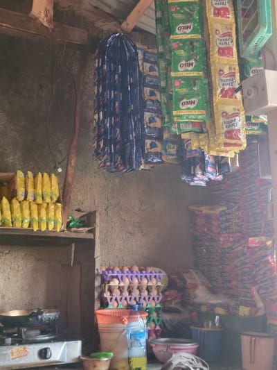 The image depicts the interior of a small convenience store in Madalla, Nigeria. The store appears to be cluttered, with shelves and hanging displays stocked with various food items and beverages. In the foreground, a gas stove with a pan on it suggests food preparation. To the right, stacks of packaged goods line the walls, some of which are identifiable as Milo and Three Crowns products, common brands in Nigeria. An egg carton filled with eggs is also visible, along with buckets and bottles of water. The store's walls are unplastered, revealing a rough concrete or mud texture. The ceiling is supported by wooden beams. The overall impression is of a modestly stocked local shop. There are no people visible in the image.