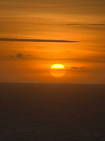 This image captures a sunset over the ocean, viewed from the vicinity of Pecatu, Indonesia. The upper two-thirds of the vertical frame are dominated by a sky displaying a gradient of intense orange, yellow, and reddish hues, indicative of late twilight. Multiple horizontal cloud formations, varying from thin, streaky cirrus-like structures to more diffuse patches, are visible against the illuminated sky. The sun, a prominent and intensely bright circular disc colored vivid yellow and orange, is centrally positioned just above the indistinct horizon line, appearing partially obscured by atmospheric haze. The lower third of the frame depicts the vast expanse of the ocean, rendered in dark, muted orange-brown and grey tones, reflecting the diminished light. The water surface appears calm with minimal discernible texture. No individuals, man-made objects, or specific actions are present or identifiable within the scene.