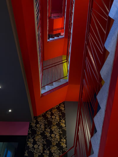 The image depicts a dramatic, brightly colored spiral staircase viewed from above. The staircase features vibrant red railings and an orange wall. The steps are made of a textured material, possibly concrete or stone, and are illuminated from above. The surrounding walls are a mix of dark gray, orange, and a patterned wallpaper with floral motifs in yellow and black. The overall impression is one of modern, bold architectural design. The location is the staircase of the Städel Museum in Frankfurt, Germany. There are no people visible in the image.