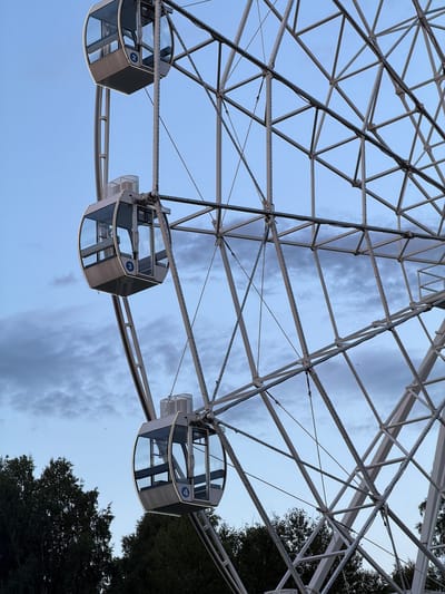 This is a low-angle, rotated photograph of a Ferris wheel in Nizhnekamsk, Russia. The subjects are the gondolas of the Ferris wheel, which are enclosed, hexagonal structures with glass windows and metal framing. There are at least four gondolas visible in the image, numbered 2, 3, and 4. The Ferris wheel's metallic structure is intricate, with a lattice of beams and supports against a cloudy sky. Trees are visible on the right side of the image, suggesting an outdoor setting, possibly a park or amusement area. The lighting suggests it is either dawn or dusk, with a soft, diffused light. The sky is a pale blue with streaks of grey clouds, indicating overcast or partly cloudy weather. The overall mood is calm and serene, with the Ferris wheel appearing stationary. There are no people visible in the gondolas. The image captures the scale and geometric complexity of the Ferris wheel's construction.