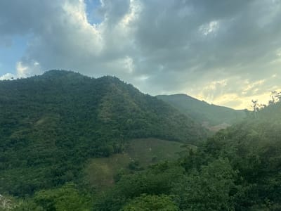 The image shows a panoramic view of lush green mountains under a cloudy sky. The terrain is rugged and covered with dense vegetation, with some areas showing signs of cultivation or clearing, such as terraced fields on the slopes. The sky is dramatic, with large, gray clouds interspersed with patches of lighter sky and hints of sunlight breaking through. The overall impression is one of natural beauty and a rural landscape, likely in Aê Thi, Vietnam, given the context. There are no people or specific objects clearly visible in the scene besides the natural environment itself.