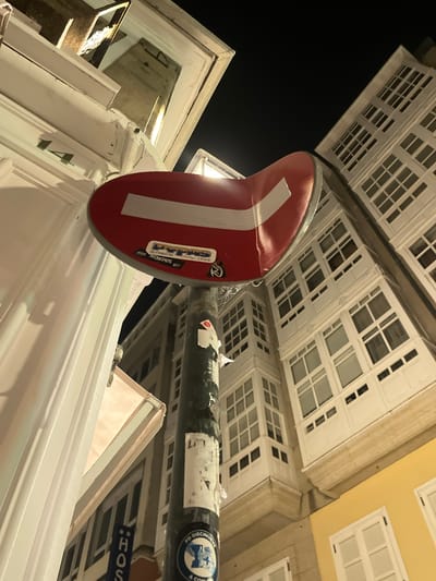 The image, captured at night, presents a low-angle view of a no-entry traffic sign mounted on a dark, cylindrical pole. The sign is red with a horizontal white bar. Several stickers adorn the sign, including one with text that appears to read "LAVIPAS" and another with "TORCAS." The pole itself is weathered and covered in various papers and stickers, one circular sticker at the bottom has a blue border with an emblem and text that seems to read "PD DIECINUEVE A COMU."
Behind the sign, a white building with numerous windows is visible, characteristic of the architecture found in A Coruña, Spain. The windows are framed in white and set into the building's facade, which is also predominantly white. The building's design, with its enclosed balconies or "galerías," is a distinctive feature of this Galician city. To the right of this building, another structure with a bright yellow facade is partially visible, also featuring white-framed windows.
The overall setting suggests an urban environment, likely a street in A Coruña, given the architectural style. The lighting indicates it is nighttime, with artificial lights illuminating the buildings and the street sign. The perspective is tilted upwards, making the buildings appear to loom over the viewer.
