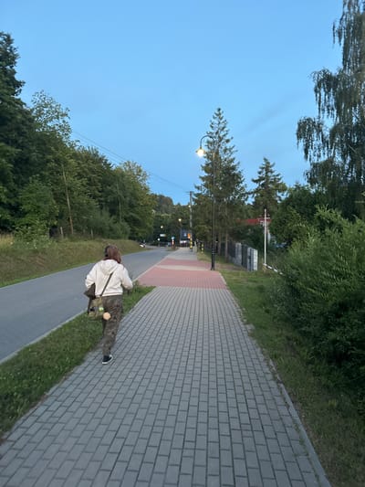 A person with long brown hair, wearing headphones, a white hoodie, and camouflage pants, walks away from the camera down a brick sidewalk next to a paved road. They are carrying a crossbody bag. The scene is an outdoor environment with trees and vegetation on the left and right sides of the road and sidewalk. A streetlamp is visible in the background. The sky is a clear blue. The location is identified as Chmielno, Poland.