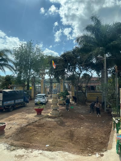 A bright, sunny day illuminates a temple complex. Ornate pillars and fences line a dirt path where people stroll. A flag with colorful stripes hangs from a tree. A truck and a white van park within the grounds. Lush trees, including a palm, frame the scene. A toy airplane hangs from a tree branch against a backdrop of blue sky with puffy clouds.