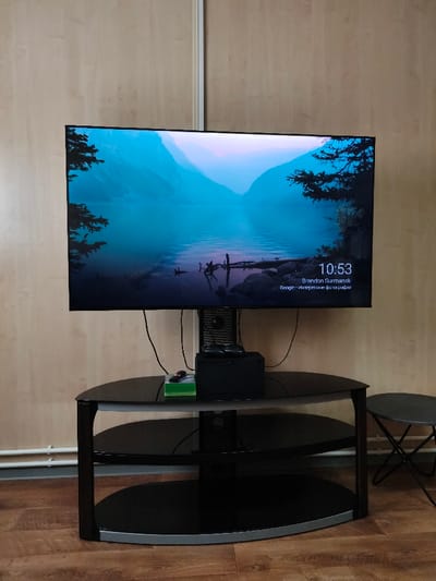 An indoor scene in the city of Im. Gastello, Russia, features a television and media setup against a light wood-paneled wall. A large, black-framed flat-screen television is centrally positioned, displaying a digital image of a serene lake bordered by mountains and evergreen trees, rendered predominantly in shades of blue and purple. The television screen shows "10:53" in the bottom right corner, along with text identifying "Brandon Surmanski" and "Google - интересные фотографии".

Below the television, a three-tiered media stand with dark glass shelves and a dark frame with silver base trim is situated on a light brown wooden laminate floor. On the top shelf of this stand, a black rectangular electronic device is positioned, topped by a smaller black device, and to its left, a translucent green rectangular container is present. Multiple black cables descend from behind the television and the devices on the stand. To the right of the media stand, a small, dark gray round stool with black folding legs is visible. In the lower left corner of the scene, two white horizontal heating pipes are mounted against the wall. The walls exhibit a vertical seam or trim line running behind the television setup, with the wall section to the right showing a slightly different, lighter texture. No people are present in the image.