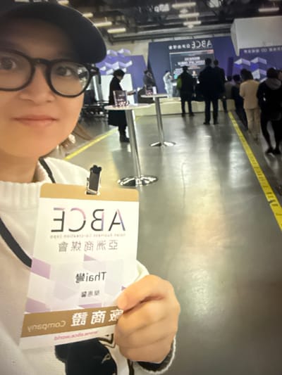 The image shows a close-up of a person wearing glasses and a baseball cap, holding up an identification badge. The badge displays text and logos, including the letters "ABCE" in large font. Below this, there are lines of Chinese characters and a smaller English website address. In the background, a large conference or exhibition hall is visible, with several people standing around tables and a large screen displaying text and graphics. The environment appears to be indoors, with a polished floor and ceiling lights. The overall scene suggests a business or technology event. The specific location in Taipei, Taiwan, cannot be determined from the visual information provided.