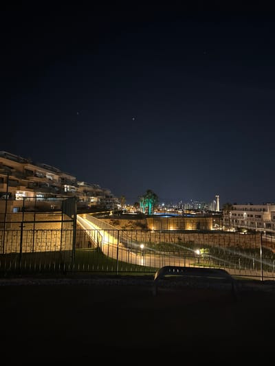 A nighttime exterior shot captures a tiered residential complex built into a hillside in Finestrat, Spain. The buildings, with their multiple balconies and lights, are illuminated, creating a striking contrast against the dark sky. A lit pedestrian bridge crosses an area with stone walls and landscaping, leading towards a pool and more buildings in the distance. Palm trees are visible, one of which is illuminated with green lighting. The city skyline of Benidorm can be seen in the background. A metal fence runs along the foreground, and a dark, modern bench sits on the pavement. The sky is clear and dark, with a few faint lights visible.