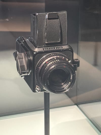 This is a close-up shot of a vintage Hasselblad camera, a high-end medium format camera known for its professional quality and durability. The camera is black with a textured leatherette grip on the film back. Attached to the body is a Carl Zeiss Planar 1:2.8 f=80mm lens. The lens has various rings for aperture and focus control, with markings visible. The serial number "Nr6196007" is clearly printed on the lens.
The camera appears to be displayed in a museum or exhibition setting, possibly behind glass, given the reflections on the surface of the lens and body. The background is out of focus, showing a muted color palette with hints of grey and white, suggesting an indoor, controlled environment. The lighting is diffuse, highlighting the details of the camera. There is no person visible in the frame. The scene is static, focusing solely on the camera as an object of display. The setting is identifiable as Rotterdam, Netherlands, though no specific landmarks are visible in the immediate vicinity of the camera. The overall impression is one of a well-preserved piece of photographic history.