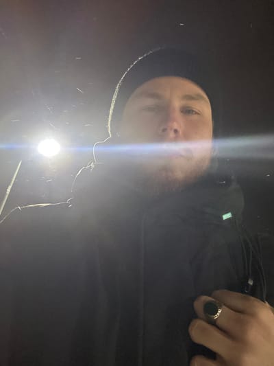A man's face is visible from a low angle, illuminated by a bright light from below, causing overexposure. He is wearing a dark hat and jacket. A ring with a large, dark stone adorns his right hand. The lighting creates stark shadows and obscures some details, making it difficult to discern the exact surroundings or specific features. The image composition is somewhat unconventional, focusing on the upward perspective.