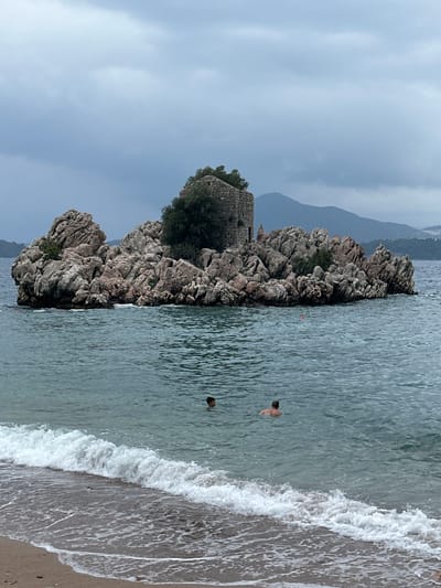 A serene coastal scene unfolds in Pržno, Montenegro, under an overcast sky. The image features a prominent rocky outcrop or small island on the left side, covered in rugged, greyish-brown rocks and sparse green vegetation, with trees at its peak. Perched atop this rocky formation, partially obscured by the foliage, are the weathered ruins of an old stone structure, possibly a tower or fort.
To the right of the rocky formation, the calm, greenish-blue sea extends, showing gentle ripples on its surface. Further to the right, a sandy beach meets the water, where soft, white-foamed waves are breaking along the shore. Two individuals are visible in the water, apparently swimming or wading. One person is closer to the beach, with their head and upper back visible, while another is further out, only their head showing above the waterline.
In the background, distant mountains rise under the diffused light of the cloudy sky. The overall impression is one of peaceful leisure on a calm, perhaps late afternoon or early evening, cloudy day. There is no visible text in the image.