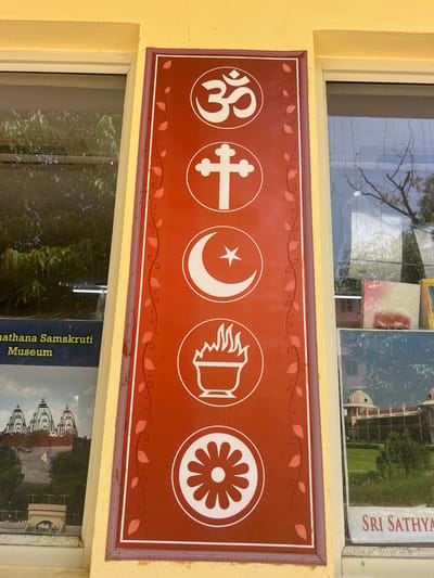 The image displays a vertical red sign with several religious symbols depicted in white circles. From top to bottom, the symbols are Om (Hinduism), a Christian cross, a crescent moon and star (Islam), a bowl with flames (Zoroastrianism), and a Dharmachakra (Buddhism). The sign is bordered by a dark purple and light purple floral pattern. To the left of the sign is a window with text that reads "Snathana Samskruti Museum". To the right of the sign is another window showcasing images of buildings, one of which has the text "SRI SATHYA" visible. The building is typical of the architecture found in Puttaparthi, India, a town known for its spiritual significance and the ashram of Sathya Sai Baba. The yellow painted wall frames the entire scene, suggesting the exterior of a building in Puttaparthi.