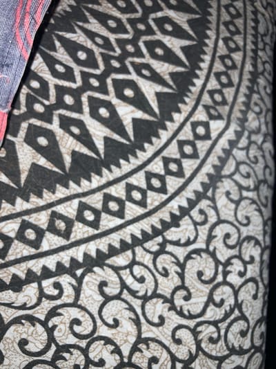 The image shows a close-up, angled view of a fabric with a detailed pattern. The pattern features geometric shapes, including diamond-like forms with circles in their centers, arranged in a band. This band is bordered by jagged, triangular shapes on one side and a more intricate, swirling, vine-like pattern on the other. The background fabric has a subtle, lighter paisley or floral print. On the upper left side of the image, a portion of another fabric is visible, appearing to be denim or a similar material with thin pink stripes.
The image does not contain any information about the city of Sabadell, Spain, or any people, actions, or interactions. It is solely a close-up of a textile.