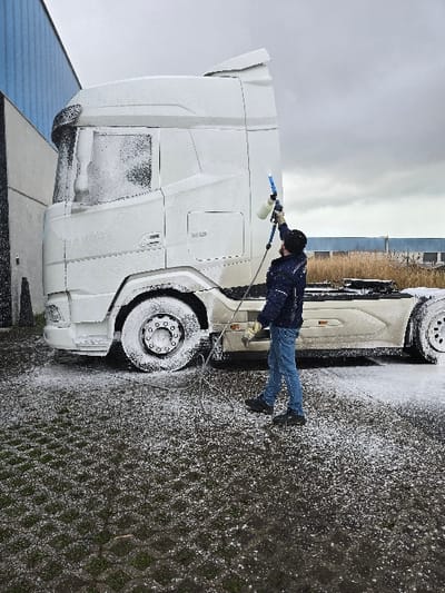 A single individual is observed applying a thick white foam to a large white semi-truck in an outdoor location in Ostend, Belgium. The person, dressed in a dark blue jacket, blue jeans, and dark shoes, holds a blue and white spray lance with an attached foam reservoir, directing the stream towards the truck's cabin. The truck's cabin, chassis, and wheels are largely covered in a dense layer of white foam. The ground beneath the truck and person consists of dark gray-green permeable pavers, wet and partially coated with foam. In the immediate background to the left, a large industrial building with blue corrugated siding is visible. Further in the distance to the right, an area of dry reeds and a building with a blue roofline are present under an overcast gray sky.