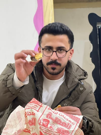A male individual, appearing to be in his late twenties or early thirties, with dark hair, a moustache, and black-rimmed glasses, is positioned centrally in an indoor setting. He is wearing a dark olive-green puffer jacket over a white long-sleeved shirt and a dark watch on his left wrist. He looks directly forward. His right hand is extended, holding a partially consumed small sandwich or pastry with a light-colored bun and a dark filling. His left hand is holding a white paper wrapper adorned with red Arabic text and graphics, indicating "معجنات أبيض المحلية" (Local White Pastries) and listing specific Damascus locations and phone numbers. The background consists of a light-colored wall featuring a vertical pink wavy decorative element and a vertical yellowish-beige strip. The scene is located in the city of Al-Qanawat Municipality, Syria.