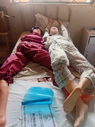This image depicts a medical training simulation in a room at D.E.C. College of Nursing, located in Ochadamu, Ofu Local Government Area, Ejule, Kogi State, Nigeria.

Two full-body medical training mannequins are positioned supine on a single bed with a white metal frame. The mannequin on the left wears a maroon pajama set; its head rests on a pillow, and its lower legs and feet are exposed prosthetic components. The mannequin on the right wears a cream-colored, patterned pajama set; its head rests on a pillow, and both arms are extended upwards above its head. Its lower legs and feet are also exposed prosthetic components.

On the bed, between the mannequins' lower bodies, a blue plastic sheet partially covers a document titled "NURSING PROCEDURE MANUAL," which is further identified with the institution's name and a visible year "2022." The bed is covered with light-colored sheets. To the immediate left of the bed, a partial view of a wooden cabinet or shelf is visible. To the right of the bed, a light-colored wall features faint markings, and an overhead window with horizontal security bars allows ambient light into the room.