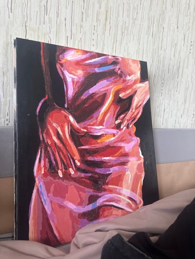 The image shows an acrylic painting on a black canvas. The painting depicts two figures intertwined, with a focus on hands and bodies. The color palette is dominated by shades of red, pink, and purple, with some orange and blue accents. The figures appear to be embracing. The painting is sitting on a surface that includes a light gray, textured pattern and a darker gray, plain area. A piece of fabric, possibly clothing, is visible in the background on the right side of the image. The location of Высокий, Russia is not determinable from the information presented in the image.