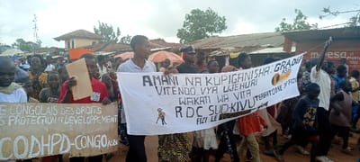 A crowd of approximately 30-50 individuals, including adults and numerous children, are engaged in a street procession within the city of Fizi, Democratic Republic of the Congo. The procession is moving forward on an unpaved, dirt ground.

Central to the scene is a large white banner, approximately 3-4 meters in length, held horizontally by at least five individuals. The banner features prominent black text, including "AMANI NA KUPIGANISHA VITENDO VYA UJEHURI WAKATI WA VITA RDC SUD-KIVU FIZI LE [date partially obscured]" and "GRADEF ASBL" with a circular logo, and "TERRITOIRE DE FIZI." An illustration of a stylized human figure is present on the left side of this banner.

To the left of the main banner, another rectangular sign, light brown in color, is held upright. Visible black text on this sign includes "E CONGOLAIS DES DROITS," "PROTECTION ET DEVELOPPEMENT," and "CODHPD-CONGO ASBL." An individual in a red shirt directly behind this sign holds a plain brown piece of paper or cardboard. Another individual on the far right holds a thin, light-colored pole vertically.

The participants are dressed in varied civilian attire. In the background, several single-story structures with corrugated metal roofs are visible, along with a more substantial two-story building featuring a red-tiled roof and light-colored walls in the mid-distance. An orange market umbrella is partially visible behind the crowd on the left side. A tall metal lattice structure, possibly a communication tower, stands on the far left. Green trees and foliage are present in the background. A shop front displaying the partially visible word "mesa" is on the far right. The sky is overcast.