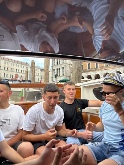 The image captures a group of young people on a boat in Venice, Italy. The boat appears to be passing along a canal, with the architecture of Venice visible in the background, including buildings with arched windows and ornate facades.
The group consists of at least five individuals. One person is wearing a striped "VENEZIA" cap and sunglasses. Several others are wearing light-colored t-shirts; one appears to be wearing a black t-shirt. The boat is moving through the water, and the reflections of the people and surrounding buildings are visible on a glass surface, likely a boat window or partition. One person can be seen steering another boat. The group seems to be looking at something in front of them, gesturing with their hands.
