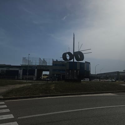 In Girona, Spain, a contemporary building complex features a striking metal sculpture composed of two large circular rings. Above the main structure, a fenced-off area, possibly a sports court, has several people visible. The building has multiple windows and signage including "PADEL PERELLO" and "OBEN." In the foreground, a crosswalk is marked on an asphalt road, with a grassy verge between the road and the buildings. Several cars are parked in front of the buildings, and a white van is also present. The sky is clear with a few wispy clouds, and a contrail from an aircraft is visible. The lighting suggests it is daytime.