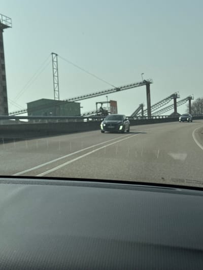 The image displays a forward view from within a vehicle, capturing an industrial landscape in Altenheim, Germany, specifically a gravel and sand processing plant likely located near Hauptstraße 55, 77743 Neuried-Altenheim. The vehicle's dark gray, textured dashboard occupies the lower portion of the frame. An asphalt road, delineated by white double solid lines and a partially visible white directional arrow in the right lane, extends into the mid-ground. The sky above is clear and light blue. Approaching on the opposing lane is a dark-colored hatchback, displaying illuminated daytime running lights. A second, less distinct dark-colored vehicle follows at a greater distance. The background is dominated by the industrial complex, characterized by multiple elevated metal conveyor belts supported by tall lattice structures. These belts connect various processing units, including a prominent green building or hopper and a tall, slender tower. A reddish-orange sign bearing the "UH" logo is affixed to one of the industrial structures. Bare-branched trees are visible on the far right horizon. No individuals are present in the scene.