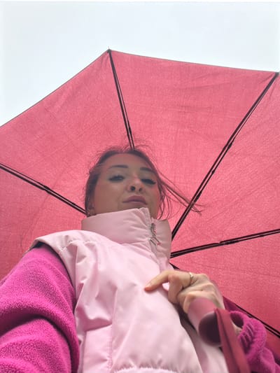 A woman, partially obscured by a pink umbrella, is looking up towards the camera. She is wearing a pink puffer vest over a fuchsia fleece jacket. Her left hand is resting on the puffer vest, and she is wearing a ring on her index finger. The background consists of the pink umbrella and an overcast sky. The location of this image is a city in Belebei, Russia.