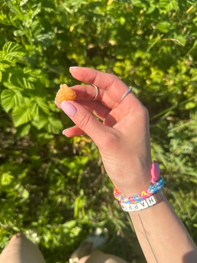 A person's hand, with manicured nails and silver rings, holds a single, pale yellow raspberry against a backdrop of lush green foliage. The hand is adorned with a colorful woven bracelet with a pink butterfly charm and a second bracelet made of white square beads spelling out "HAPPY." The background suggests an outdoor, natural setting with sunlight dappling the leaves. The image does not provide any information regarding the city of Turovo, Russia, or any specific urban location.