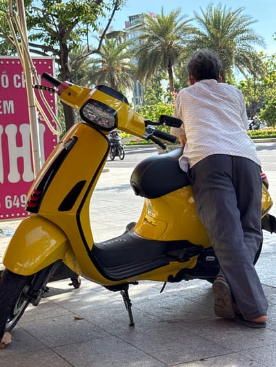 This media file shows a person dressed in a white polka-dot shirt and dark grey pants, leaning over a bright yellow Vespa scooter. The person's back is to the camera, and they appear to be interacting with the scooter. Their hair is short and grey. They are wearing dark sandals. The scooter is parked on a sidewalk, with a grey tiled ground. In the background, there are palm trees and lush green foliage, suggesting a tropical or subtropical environment. The sky is visible and appears clear. A pink sign with white lettering and pink dots is partially visible in the lower left corner. Some of the visible text on the sign appears to be "O CH" and "EM" and "649". The scene suggests a daytime setting, possibly morning or afternoon, given the bright sunlight casting shadows. The presence of palm trees and the style of the scooter hint at a location in a warm climate, consistent with the provided location context of Quy Nhon, Vietnam. The overall mood is casual and relaxed.