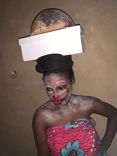 In Agban, Nigeria, a person with dark skin is posed against a plain, earthen-colored wall. They are wearing an elaborate face paint design, with white powder covering their face and chest, bright pink eye shadow, and dark red lipstick applied in a clown-like fashion. A black headwrap is worn, and over this, a white box is balanced, topped by a round, textured object resembling a gourd or a weathered bowl. The individual is draped in a brightly colored fabric featuring a pink background with blue floral patterns and black lines. The lighting is dim, creating shadows and highlighting the facial makeup. The setting appears to be indoors, possibly a dwelling, given the wall texture and the indoor nature of the objects.