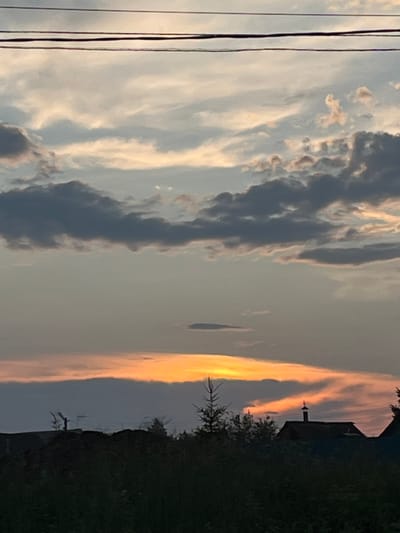 This is a vertical, medium-range photograph of a sunset over a rural area, likely in Ludorvay, Russia. The sky dominates the upper two-thirds of the frame, filled with a mix of grey, white, and orange clouds. Three thin, dark power lines stretch horizontally across the top of the image. The lower third of the image shows a silhouetted landscape of trees and rooftops against the brightly lit sky. One prominent building has a chimney with a small, pointed cap. A thin, spiky evergreen tree stands to the left of the building. The overall impression is one of a peaceful, twilight scene.