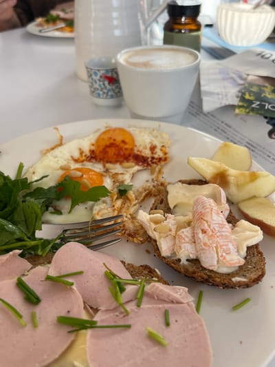 The image displays a meal set on a white table, located in a dining establishment in Waldgrehweiler, Germany. In the foreground, a white oval plate holds a variety of food items. These include two fried eggs with distinct orange yolks, one of which shows signs of partial consumption, seasoned with red spice and garnished with pine nuts and green herbs. A metal fork rests on the plate. Alongside the eggs are a portion of green leafy vegetables and several slices of red-skinned apple, one exhibiting a visible bite mark. Two slices of dark, seeded bread are also on the plate; one is topped with light pink, thinly sliced cold cuts and scattered green chives, while the other features a white spread layered with a reddish-orange soft cheese-like substance, garnished with pine nuts. In the background, on the white table, a white ceramic pitcher, a small blue and white patterned ceramic cup, and a white cup containing a foamed beverage are visible. Further back, an amber bottle with a dark lid, a small white ribbed bowl, and printed material with partial text are present. A blurred human figure occupies the far background.