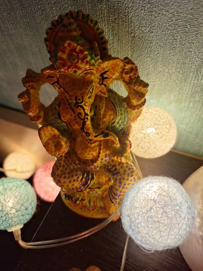 A close-up, high-angle shot shows a colorful, ornate statue of Ganesha, a Hindu deity, positioned in the center of the frame. The statue is yellow with red, green, blue, and purple painted accents and intricate patterns. It appears to be made of resin or a similar material. Surrounding the statue are several round, spherical decorative lights, commonly known as cotton ball lights or fairy lights. Two lights are visible in the foreground on the left, one in a light blue hue and another in pink. To the right and behind the statue, a larger, cream-colored light glows brightly. Another light, also cream-colored, is partially visible in the far right background. The background surface behind the statue is a textured, light-colored wall, possibly wallpaper or a painted surface with a patterned finish. The statue and lights rest on a dark, flat surface, likely a table or shelf. The lighting in the image is soft and ambient, with the fairy lights providing the primary illumination, creating a warm and decorative atmosphere. The image does not contain any elements that would allow for the determination of its location within the city of Vologda, Russia.