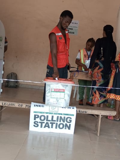 The scene depicts a polling station located in Ajalli, Nigeria. In the foreground, a transparent ballot box sits on a wooden bench, containing numerous folded white papers. A white sign with green lettering, "INDEPENDENT NATIONAL ELECTORAL COMMISSION POLLING STATION," is placed on the floor in front of the bench. A dark-complexioned male, wearing a red sleeveless vest and dark trousers, stands over the ballot box, holding a white cylindrical object, appearing to deposit an item. His gaze is directed downward.
Behind the ballot box, a dark-complexioned female with dreadlocks, wearing a white shirt and an orange safety vest, stands behind a small wooden table, attending to materials. To her right, another dark-complexioned female with long braided hair, dressed in a black top and a floral patterned skirt, stands partially visible. A white string or rope stretches horizontally across the lower mid-ground.
The background features a light-brown wall with election-related posters, including "EC 30 C" and "VOTING IN PROGRESS." To the far left, part of a white voting booth is visible. A grey backpack lies on the tiled floor near the voting booth. The floor is tiled with light-colored square tiles. A water bottle is visible on the floor to the right. The wooden bench has "LEGION OF MA" and "UKWU" inscribed on it.