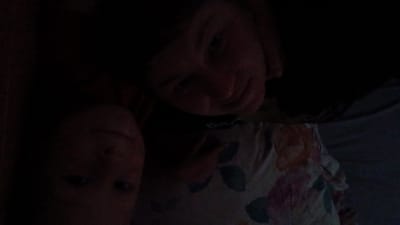In a dimly lit room, two individuals are captured in a selfie, positioned close to the camera. The person in the foreground is lying down, with their head tilted back, and their face is partially obscured by shadows. They appear to be wearing a shirt with a floral pattern. The second individual is partially visible behind the first, their face also in shadow. The overall scene suggests an intimate, informal setting, possibly a bedroom or living space. The exact location within the city of Паренда, Russia, is not discernible from the image.