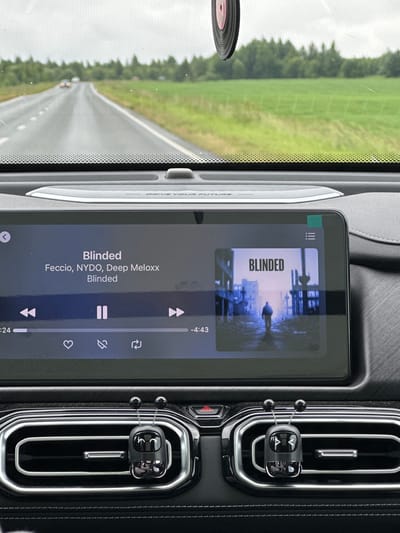 The image shows the interior of a car, viewed from the driver's perspective. A large touchscreen display is centrally located on the dashboard, showing the album art and title "BLINDED" for a song by Feccio, NYDO, and Deep Meloxx. The song is currently playing, with the playback time displayed as 4:43. Below the song information, there are icons for liking, adding to a queue, and shuffling the playlist. The car is on a paved road, with a green field and trees visible through the windshield. The sky is overcast. Two car air fresheners, shaped like small black robots with antennae, are attached to the car's air vents. The car appears to be in motion, suggesting the photo was taken while driving. The location is identified as Maksimovo, Russia.