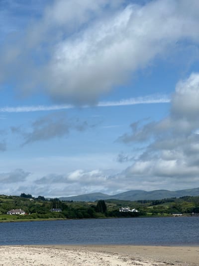 The image depicts a serene waterfront scene in Tullycullion, Ireland. In the foreground, a sandy beach is visible, with the gentle lapping of a dark blue body of water. Across the water, a lush green landscape unfolds, dotted with trees and rolling hills in the distance. Several houses, with white exteriors and dark roofs, are nestled among the trees on the shoreline. The sky above is a mix of blue and white clouds, suggesting a partly cloudy day. The overall atmosphere is one of tranquility and natural beauty.