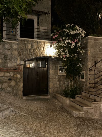 This image depicts a nighttime scene in Bartula, Montenegro, focusing on the exterior of a stone building with a wooden door and a flowering bush. The foreground is a cobblestone pathway leading up a gentle slope towards the entrance. To the left of the path, stone walls are visible, constructed from irregular, weathered stones with some visible mortar. A section of red brick is integrated into the lower part of this wall.
The main focus is a dark wooden double door set within a stone archway. The door has horizontal planks and two small, square windows near the top of each panel, offering a glimpse of interior spaces that appear to be illuminated. Above the door, a small, ornamental metal structure is attached to the stone.
To the right of the door, a blooming bush with clusters of white and pink flowers is prominently featured, illuminated by an overhead light source. Behind the bush, the stone wall continues, featuring two square window-like openings. One opening appears to contain a circular grate or vent, while the other shows a small potted plant.
Further to the right, a set of concrete stairs with a black metal railing leads upwards. The railing has decorative scrollwork at its base. The stairs are illuminated, revealing their stone steps.
In the upper left of the image, a balcony railing made of black metal bars is visible, with a small blue illuminated object, possibly a call button, attached to the stone wall below the railing. Lush green foliage from an overhead tree partially obscures the upper part of the building on the left. The overall atmosphere is one of a dimly lit, old-world charm, typical of Mediterranean coastal towns.