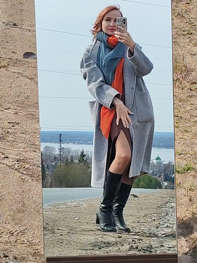 A woman is taking a selfie in a mirror. She has red hair, wears a grey coat, a brown dress with a slit, a blue and orange scarf, and black boots. The mirror is situated outdoors on a sandy surface. Reflected in the mirror is a landscape with water, trees, and a green-domed building, possibly a church. According to geolocation data, the location is in Нечкино, Russia.