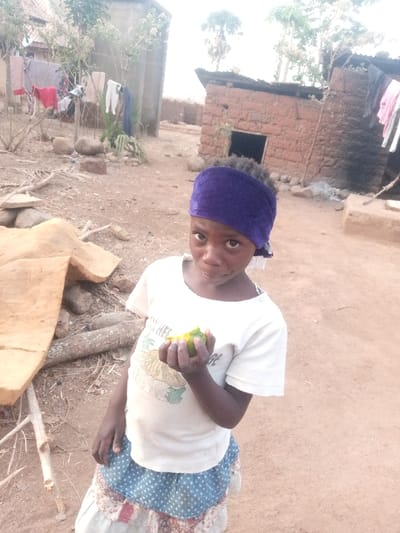 The image depicts a young child in Agban, Nigeria, standing outdoors on dry, reddish-brown dirt ground. The child, with dark skin and a neutral expression, wears a purple headwrap, a white short-sleeved t-shirt with a graphic design, and a patterned skirt. They hold a piece of green and yellow fruit in their right hand. In the background to the left, a light-colored building with a visible roofline has laundry hanging on a line or fence in front of it. To the center-right, a smaller, reddish-brown mud or brick structure with a dark doorway and a dark roof is visible, also with clothes hanging nearby. Scattered rocks, wood pieces, and sparse dry vegetation are present throughout the scene. A large, light-brown, irregularly shaped object lies on the ground to the left of the child.