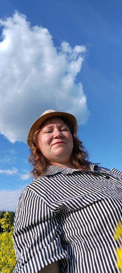 The image captures a low-angle, outdoor shot of a woman in Зипуново, Russia. The woman, positioned in the lower and central portion of the frame, has reddish-brown, wavy hair and is wearing a straw hat. Her face is directed towards the camera, exhibiting a neutral expression with a subtle smile and red lipstick. She is dressed in a black and white vertically striped button-down shirt. The background is dominated by a bright blue sky, occupying the upper two-thirds of the image, with several scattered white cumulus clouds. A prominent bright light source, likely the sun, is visible in the upper left, partially obscured by a cloud, creating a distinct luminous area. In the lower left of the image, vibrant yellow flowers are visible, suggesting a field. Faint green foliage, consistent with distant trees, is discernible on the left horizon line. Multiple thin power lines are present in the mid-ground on the left side, stretching horizontally across the sky.