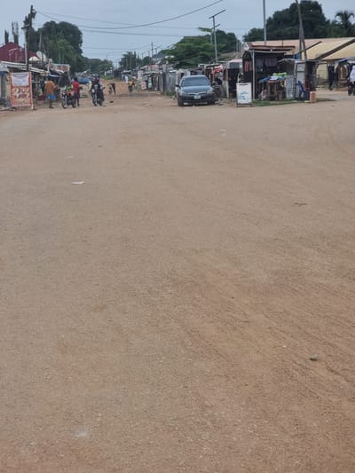 The image depicts a street scene in Gwagwalada, Nigeria, under an overcast sky. The street is unpaved and appears to be a dirt road. On either side of the road are various makeshift structures that function as shops and stalls. In the foreground, the dirt road stretches out, with scattered debris and a few pieces of paper.
To the left, several people are gathered around motorcycles. Two men are standing next to a motorcycle, while another man is seated on a motorcycle. Further to the left, a sign for "Abuja Restaurant" is visible, with images of food items. More stalls and structures are visible behind these individuals, some with goods displayed outside.
In the middle ground, a dark-colored car is parked on the right side of the road. Adjacent to the car is a small shop with goods displayed on shelves. Several people are seen near these shops, including a person in a white garment walking away from the camera. Another motorcycle is visible further down the road.
The background shows a line of trees and more buildings, suggesting a developed area within the town. The overall scene conveys a sense of everyday activity in a commercial area.