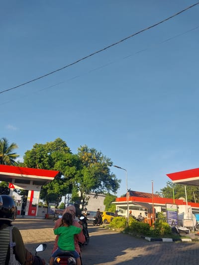 This media file depicts an outdoor scene at a gas station in Macege, Indonesia. The image is oriented vertically with the sky taking up a significant portion of the frame. Power lines are visible stretching across the blue sky.

The ground level of the scene shows a Pertamina gas station with red and white awnings. Several motorcycles are present, with people riding on them. One motorcycle in the foreground has a person in a green shirt and blue shorts, appearing to be a child, being held by another person. Another motorcycle further back shows riders wearing helmets and casual clothing. A yellow car is parked near the gas pumps.

Trees with green foliage are abundant, suggesting a tropical or subtropical environment. The time of day appears to be late afternoon or early evening, indicated by the warm, angled sunlight casting shadows. The weather seems clear and sunny.

A sign on the gas station building is partially visible and has the word "PERHATIAN" (Attention) and some smaller text, likely instructions or warnings. Another sign reads "PERTAMAX", which is a type of fuel sold at Pertamina stations.

The overall impression is a snapshot of everyday life at a local gas station, with people on motorcycles and a car, set against a backdrop of a clear sky and lush greenery.