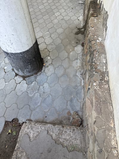 This image shows a close-up view of an outdoor scene, likely in Armavir, Russia, focusing on the ground and a concrete pillar. The ground is paved with interlocking grey concrete blocks that form a repeating pattern. The blocks appear worn and somewhat dirty, with some areas showing water stains.
Along the top of the image, there is a rough, stone-like border or wall, composed of irregularly shaped stones cemented together. This border shows signs of wear and damage, with chunks missing and loose pieces. Beneath this stone border, and above the paved ground, is a strip of dirt and debris.
A large, cylindrical concrete pillar, painted in alternating bands of white and black, dominates the lower left portion of the image. The pillar is positioned at an angle, appearing to emerge from the ground. The painted sections are weathered, with some scuff marks and areas where the paint has chipped or worn away. The pillar meets the paved ground in a slightly damaged area, with some dirt and grass growing around its base.
To the right of the paved area, there is a section of concrete that is broken and crumbling, exposing a rougher, textured surface underneath. This damaged concrete section also has dirt and some dried leaves at its base, indicating an outdoor environment.
The lighting suggests it is daytime, with diffused light and no strong shadows, possibly indicating an overcast or partially cloudy sky. The overall scene conveys a sense of age and wear, with evidence of decay and disrepair in the man-made structures. There is no visible text in the image.