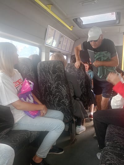 The image shows the interior of a crowded bus or shuttle van, likely in Zlatoust, Russia, given the context. Several passengers are visible, seated and standing. In the foreground, a young woman with blonde hair sits on a seat, wearing a white t-shirt and light blue jeans. She is holding a pink and purple plastic bag. Next to her, another passenger's legs are visible. In the middle ground, an older man wearing a white baseball cap, sunglasses, and a dark t-shirt is standing, holding a green bag. He appears to be interacting with someone further back. Other passengers are seated and standing in the background, with glimpses of their clothing and hair visible. The seats are upholstered with a dark fabric featuring a white leaf-like pattern. The interior of the vehicle appears functional, with overhead compartments and lighting fixtures. Natural light streams in from a window on the left, illuminating the scene. A poster or information sheet is mounted on the wall above a window. The overall impression is of a mode of public transportation during daylight hours.