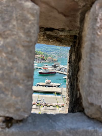 The image, framed by rough stone blocks, offers a view of a harbor in Kotor, Montenegro. The scene depicts several boats docked in clear blue water. In the background, a hillside is covered with lush green trees and dotted with buildings, suggesting a coastal town. The time of day appears to be daytime, with bright sunlight casting shadows, and the weather seems pleasant.

The boats vary in size and design, with some appearing to be smaller motorboats and others larger vessels. One boat in the foreground has the text "PORT OF KOTOR" visible on its stern. The harbor is bustling with activity, indicated by the presence of multiple boats and the dock area. The dock itself is lined with railings and planters filled with colorful flowers, adding a touch of vibrancy to the scene. The overall impression is one of a scenic and active harbor on a sunny day.